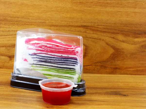 Regenbogen-Kreppkuchen in Plastikbox und süßer Erdbeersoße auf Holzgrund — Stockfoto