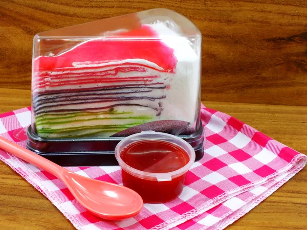 Regenbogen-Kreppkuchen in Plastikbox und süßer Erdbeersoße auf Holzgrund — Stockfoto