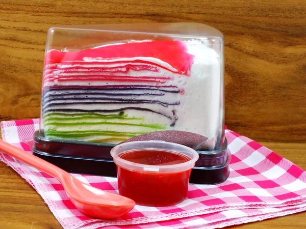 Gâteau crêpe arc-en-ciel dans une boîte en plastique et sauce aux fraises sucrée sur fond en bois — Photo