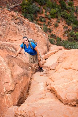 Genç bir bayan yürüyüşçü kameraya gülümsemek için kırmızı bir kayaya tırmanıyor. Sedona, Arizona 'daki popüler Katedral Rock yürüyüşünde çekildi..