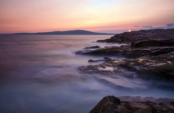 The sun sets against a rocky coast at Schoodic Point in Acadia National Park, Maine. Long exposure to achieve the misty effect of the ocean.