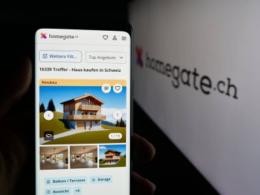 Stuttgart, Almanya - 10-09-2025: İsviçreli emlak portalı şirketi Homegate 'in web sayfası logonun önünde duran kişi. Telefon ekranının merkezine odaklan.