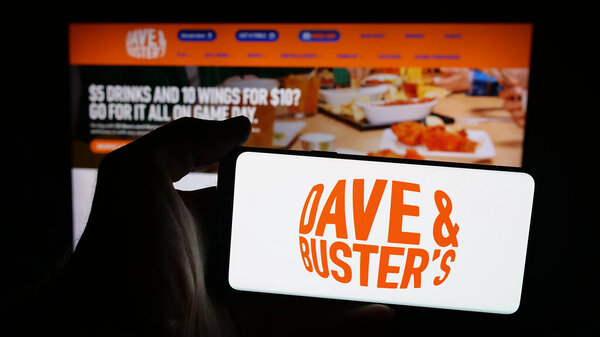 Штутгарт, Германия - 10-13-2025: Лицо, держащее мобильный телефон с логотипом компании Dave and Buster 's Entertainment Inc. на экране перед веб-страницей. Фокус на дисплее телефона.