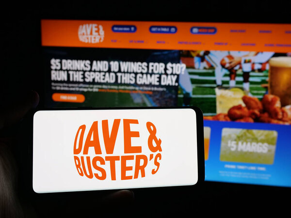 Штутгарт, Германия - 10-13-2025: Человек, держащий смартфон с логотипом компании Dave and Buster 's Entertainment Inc. на экране перед веб-сайтом. Фокус на дисплее телефона.