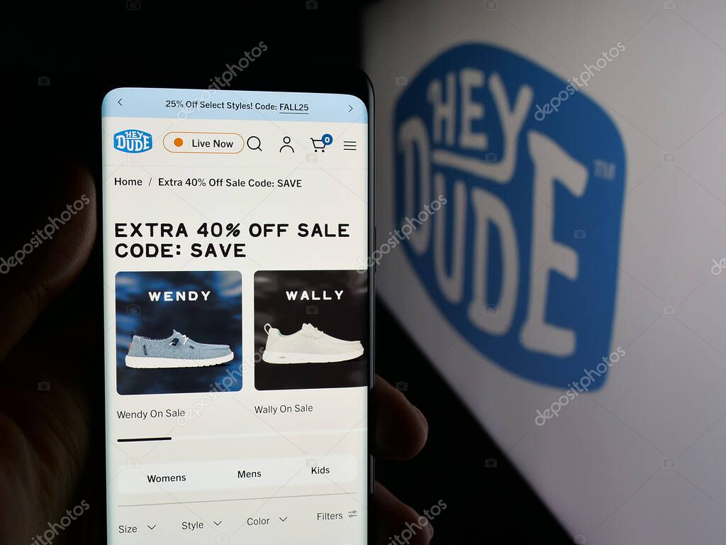 Stuttgart, Germany - 10-15-2025: Person holding cellphone with webpage of US fashion company Heydude (Heydude.com) on screen in front of logo. Focus on center of phone display.