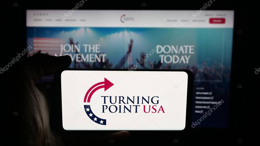 Stuttgart, Germany - 10-15-2025: Person holding cellphone with logo of US organisation Turning Point USA (TPUSA) on screen in front of webpage. Focus on phone display.