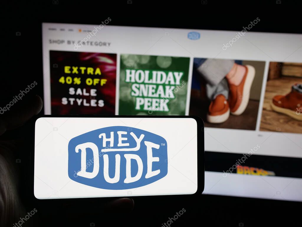 Stuttgart, Germany - 10-15-2025: Person holding smartphone with logo of US fashion company Heydude (Heydude.com) on screen in front of website. Focus on phone display.