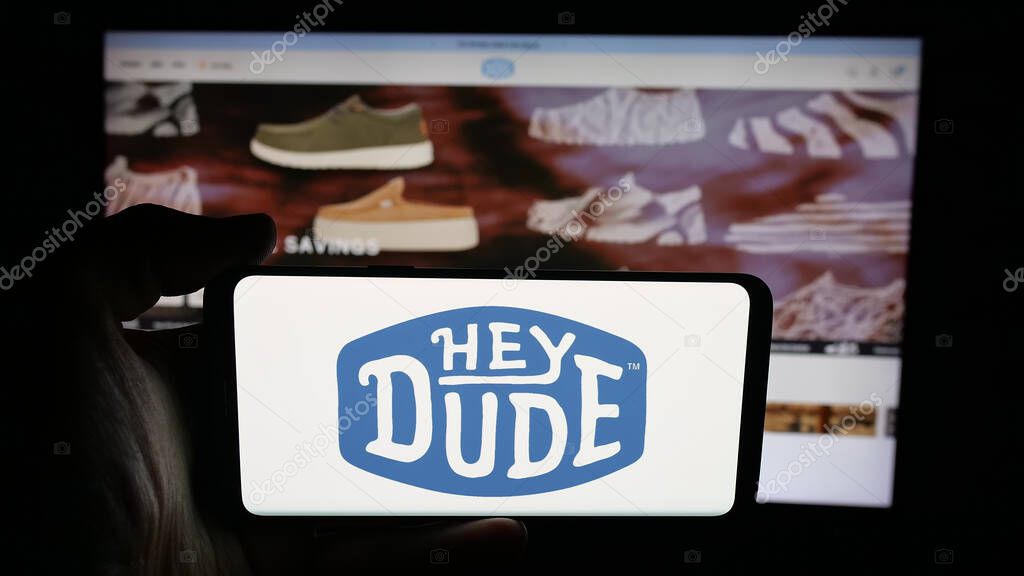 Stuttgart, Germany - 10-15-2025: Person holding cellphone with logo of US fashion company Heydude (Heydude.com) on screen in front of webpage. Focus on phone display.