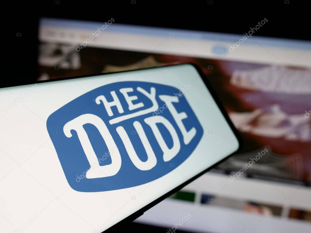 Stuttgart, Germany - 10-15-2025: Smartphone with logo of US fashion company Heydude (Heydude.com) on screen in front of website. Focus on left of phone display.