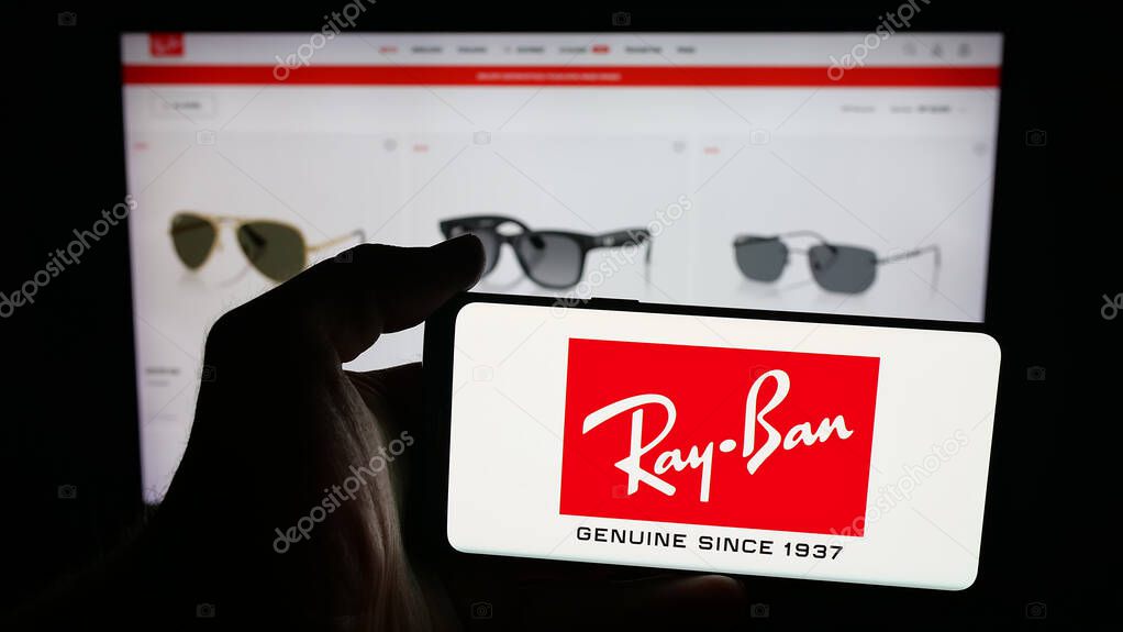 Stuttgart, Germany - 10-16-2025: Person holding cellphone with logo of luxury eyewear company Ray-Ban on screen in front of webpage. Focus on phone display.