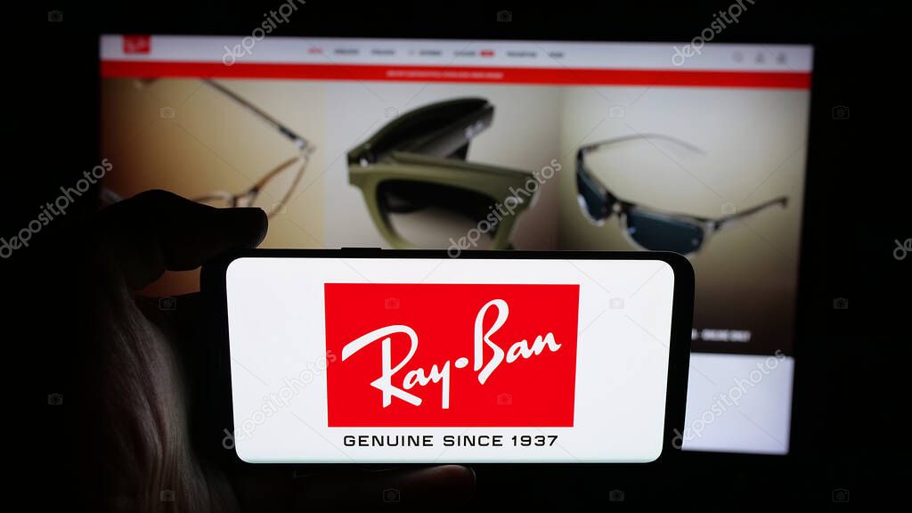 Stuttgart, Germany - 10-16-2025: Person holding mobile phone with logo of luxury eyewear company Ray-Ban on screen in front of web page. Focus on phone display.