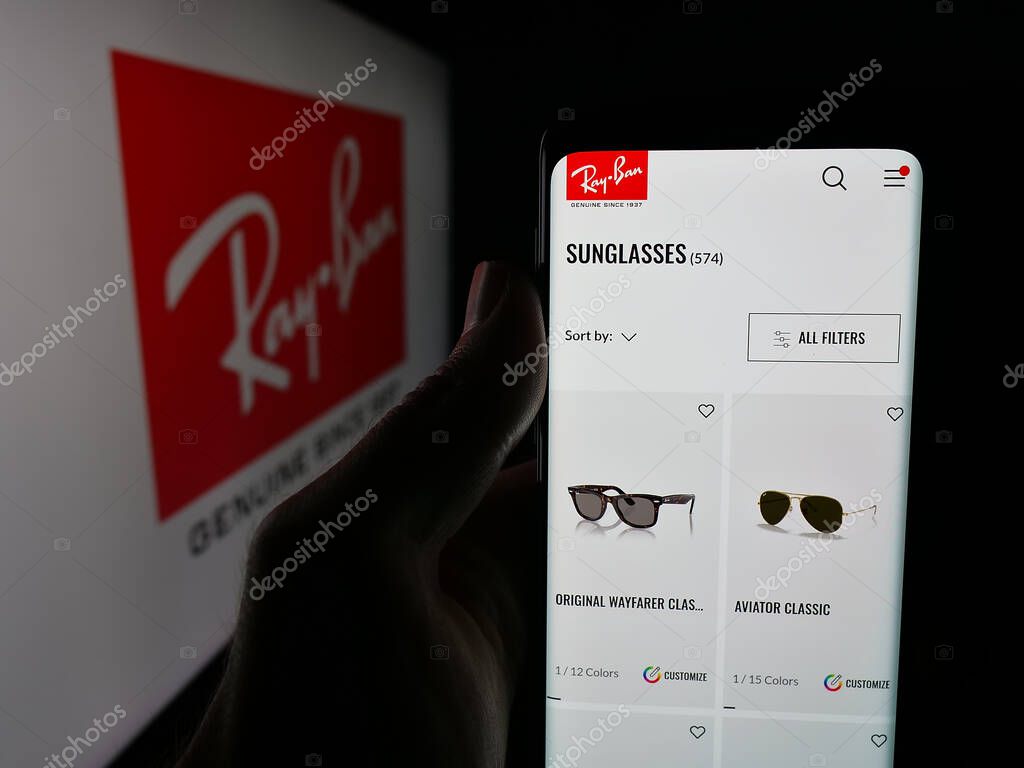 Stuttgart, Germany - 10-16-2025: Person holding cellphone with webpage of luxury eyewear company Ray-Ban on screen in front of logo. Focus on center of phone display.