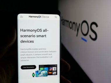 Stuttgart, Germany - 11-05-2025: Person holding smartphone with web page of operating system HarmonyOS (HMOS, Hongmeng) on screen in front of logo. Focus on center of phone display.