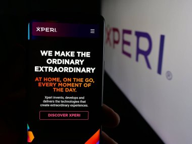 Stuttgart, Germany - 11-07-2025: Person holding cellphone with webpage of US technology company Xperi Inc. on screen in front of logo. Focus on center of phone display.