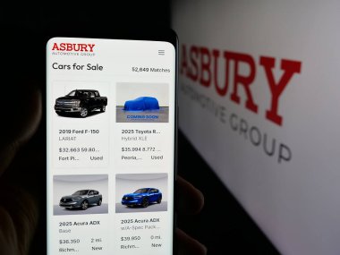 Stuttgart, Germany - 11-07-2025: Person holding cellphone with webpage of US company Asbury Automotive Group Inc. on screen in front of logo. Focus on center of phone display.