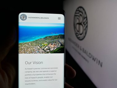 Stuttgart, Germany - 11-08-2025: Person holding cellphone with webpage of Hawaiian company Alexander Baldwin Inc. on screen in front of logo. Focus on center of phone display.