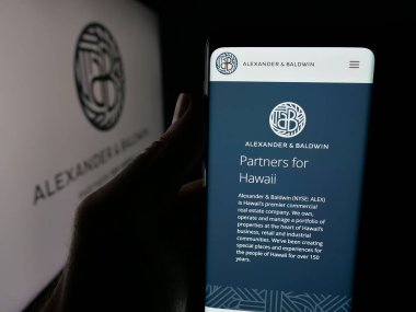 Stuttgart, Germany - 11-08-2025: Person holding smartphone with web page of Hawaiian company Alexander Baldwin Inc. on screen in front of logo. Focus on center of phone display.