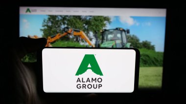 Stuttgart, Germany - 11-08-2025: Person holding cellphone with logo of vegetation equipment company Alamo Group Inc. on screen in front of webpage. Focus on phone display.
