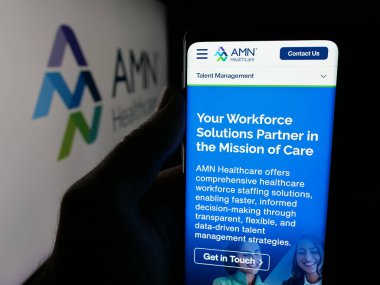 Stuttgart, Germany - 11-08-2025: Person holding cellphone with webpage of staffing company AMN Healthcare Services Inc. on screen in front of logo. Focus on center of phone display.