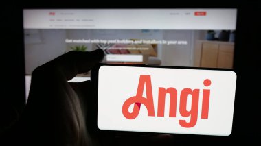 Stuttgart, Germany - 11-08-2025: Person holding mobile phone with logo of US online marketplace company Angi Inc. on screen in front of web page. Focus on phone display.