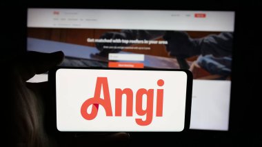 Stuttgart, Germany - 11-08-2025: Person holding cellphone with logo of US online marketplace company Angi Inc. on screen in front of webpage. Focus on phone display.