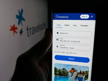Stuttgart, Almanya - 11-21-2025: ABD çevrimiçi seyahat şirketi Travelocity 'nin internet sayfası ile logo önünde cep telefonu tutan kişi. Telefon ekranının merkezine odaklan.