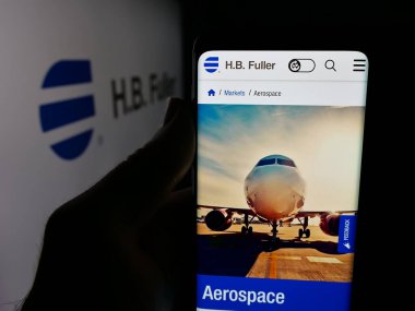 Stuttgart, Almanya - 12-19-2025: ABD 'nin yapışkan iş H.B. web sayfası ile cep telefonu tutan kişi Fuller Şirketi logonun önünde ekranda. Telefon ekranının merkezine odaklan.
