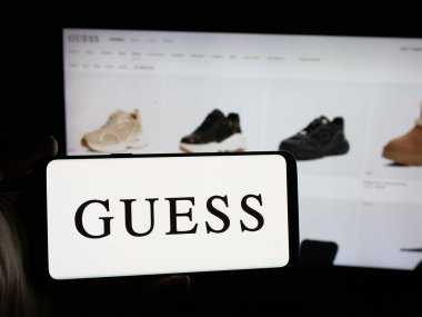 Stuttgart, Almanya - 12-19-2025: ABD 'li giyim şirketi Guess Inc.' in logosunu elinde tutan kişi web sitesinin önünde ekranda. Telefon ekranına odaklan.