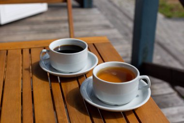 Güneşli bir sabahta evin verandasındaki ahşap bir masada bir fincan kahve ve bir fincan çay.