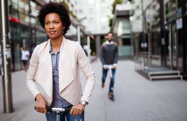 Modern iş binasının önünde elektrikli scooter kullanan iki gülen iş adamı işe devam ediyor..