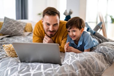 Güzel bir genç ebeveyn ve çocuk dizüstü bilgisayar kullanıyor ve evde yatakta birlikte yatarken gülümsüyorlar.