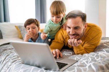 Yakışıklı baba ve çocukları dizüstü bilgisayar kullanıyorlar ve evde yatakta birlikte yatarken gülümsüyorlar.