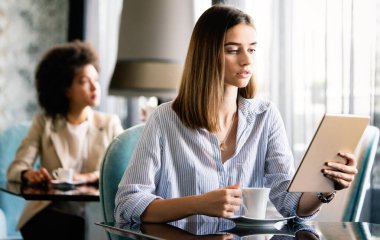 Kahve içen ve kafede tablet bilgisayar kullanan mutlu genç bir kadın.