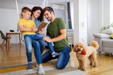 Modern aletleri olan mutlu genç bir aile ve evde oynayıp eğlenen bir köpek.