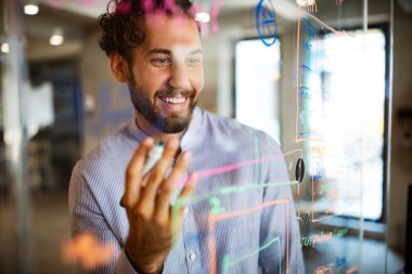 Verilerle ve diyagramlarla çalışan genç bir adam cam ofis duvarına fikirler yazıyor. İş, teknoloji, beyin fırtınası konsepti.