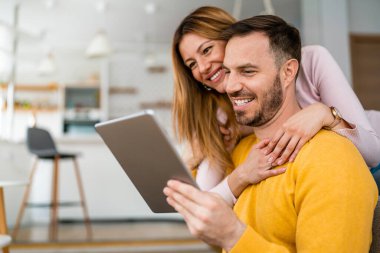 Birbirine aşık çiftler evde birbirlerine sarılıp tablet kullanıyorlar.