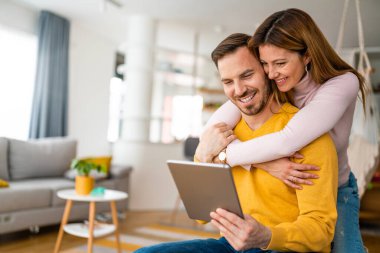 Birbirine aşık çiftler evde birbirlerine sarılıp tablet kullanıyorlar.