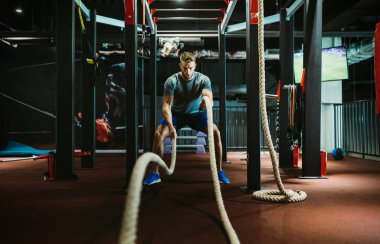 Genç formda bir adam spor salonunda savaş halatlarıyla çalışıyor.