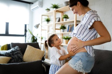 Mutlu kız çocuğu güzel hamile annenin karnına sarılıyor.