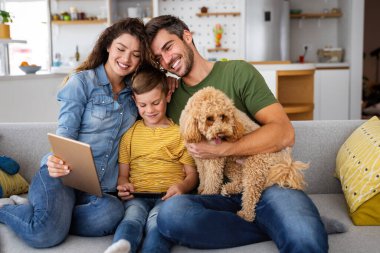 Modern aletleri olan mutlu genç bir aile ve evde oynayıp eğlenen bir köpek.
