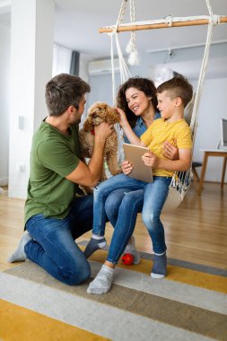 Modern aletleri olan mutlu genç bir aile ve evde oynayıp eğlenen bir köpek.