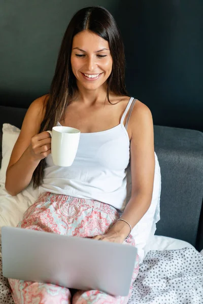 美しい女性がベッドに座ってコーヒーを片手にノートパソコンを使って — ストック写真
