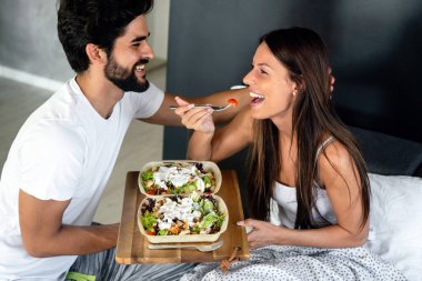 Güzel, genç, romantik bir erkek, bir kadına yatakta kahvaltı getirir ve uyandıktan sonra birlikte güler..