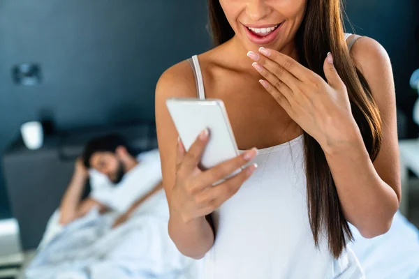 夫がベッドで寝ている間 携帯電話を使っている若い女性 浮気夫婦の概念 — ストック写真