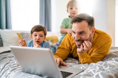 Yakışıklı baba ve çocukları dizüstü bilgisayar kullanıyorlar ve evde yatakta birlikte yatarken gülümsüyorlar.