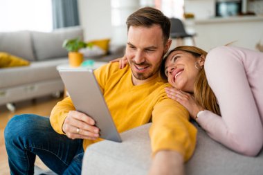 Birbirine aşık çiftler evde birbirlerine sarılıp tablet kullanıyorlar.