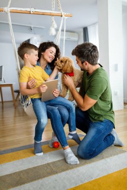 Modern aletleri olan mutlu genç bir aile ve evde oynayıp eğlenen bir köpek.