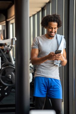 Fitness, spor, eğitim, spor, teknoloji ve yaşam tarzı konsepti. Spor salonunda akıllı telefonu ve havlusu olan genç bir adam.