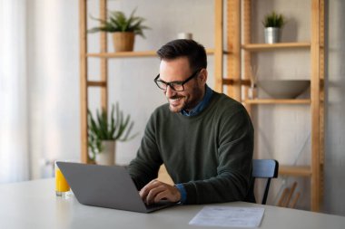 Tatmin olmuş yakışıklı adam dizüstü bilgisayarda yazıyor, internetten e-posta yazıyor, uzaktan çalışıyor ya da evde bilgisayar başında çalışıyor.