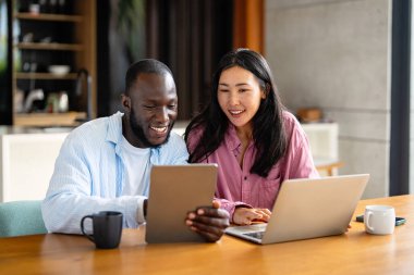 Çok uluslu çift sabah rutininin keyfini çıkarıyorlar, gülümsüyorlar ve rahat ve modern bir ev ortamında dizüstü bilgisayar ve tablet kullanıyorlar.